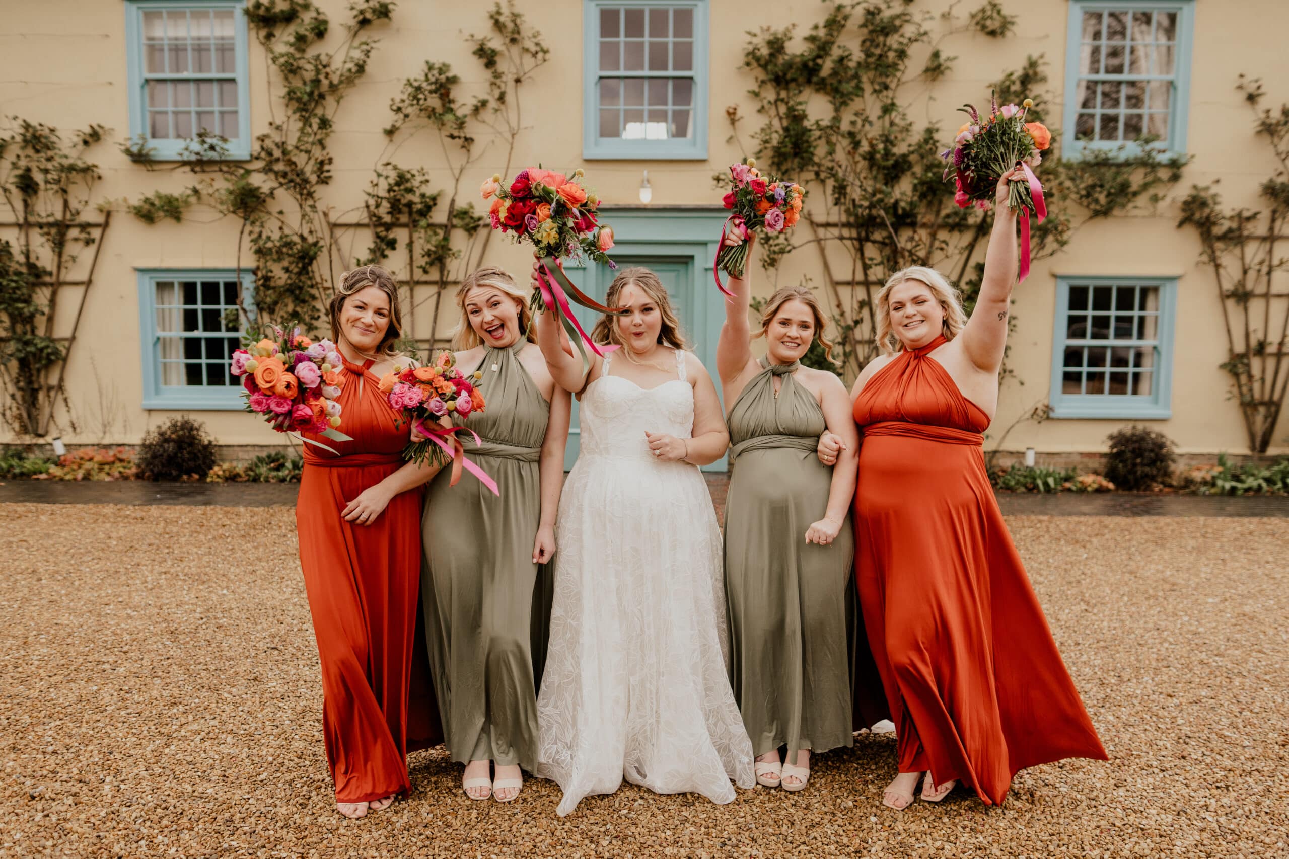 Bride in wedding dress and bridesmaids in sage green and burnt orange dresses hold colourful bouquets high in a celebratory post in front of cream country farm house with blue windows and doors