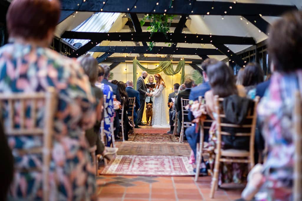 Bride and groom with their pet dog between then at barn wedding ceremony with guests seated and looking on