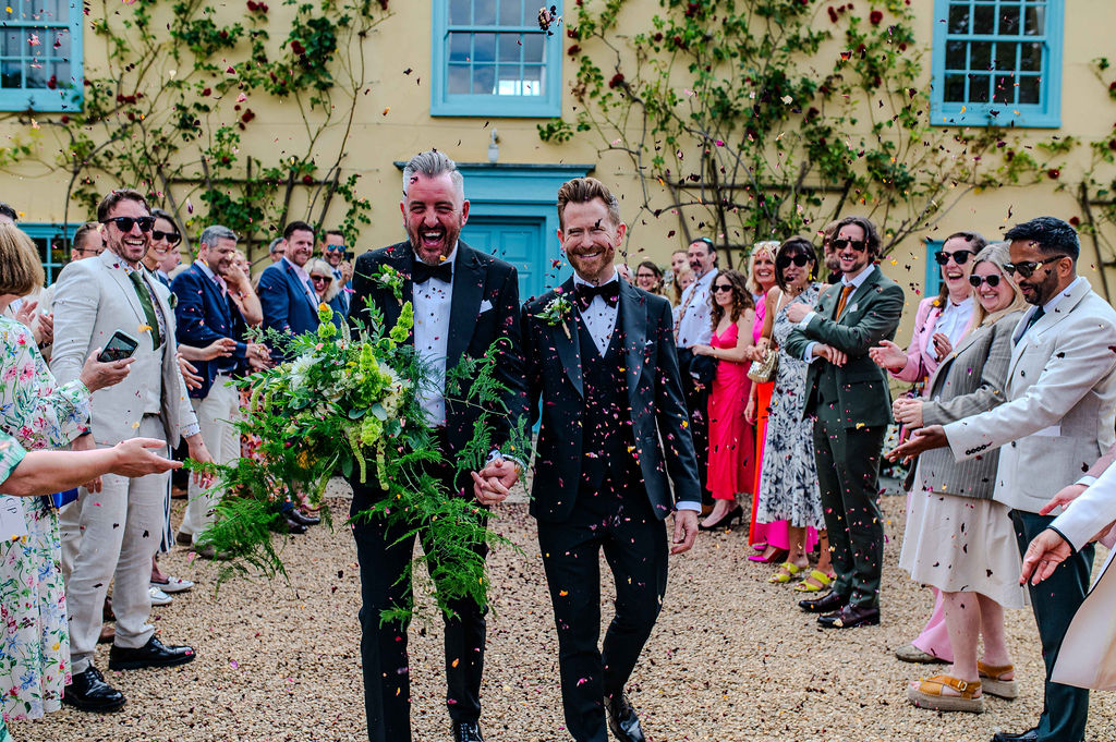 Same Sex wedding two grooms in tuxedos enjoy confetti shower from their guests in front of cream country farmhouse