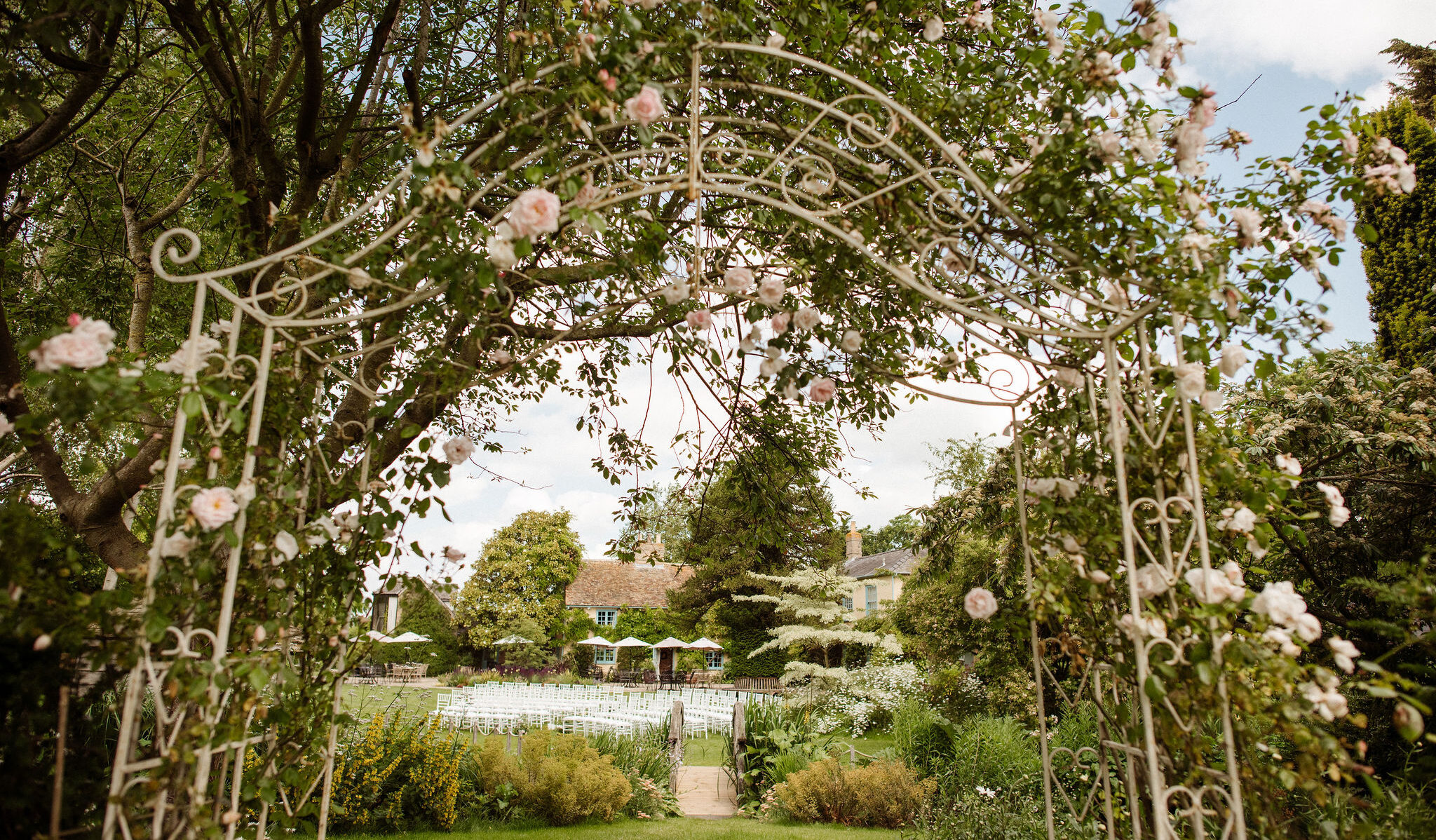 Beautiful wedding venue set for outdoor garden wedding ceremony photographed through rose pergola to the gardens and white ceremony chairs beyond