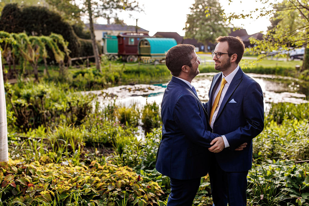 Two grooms embrace at countryside wedding with pond and country views in the backgroup