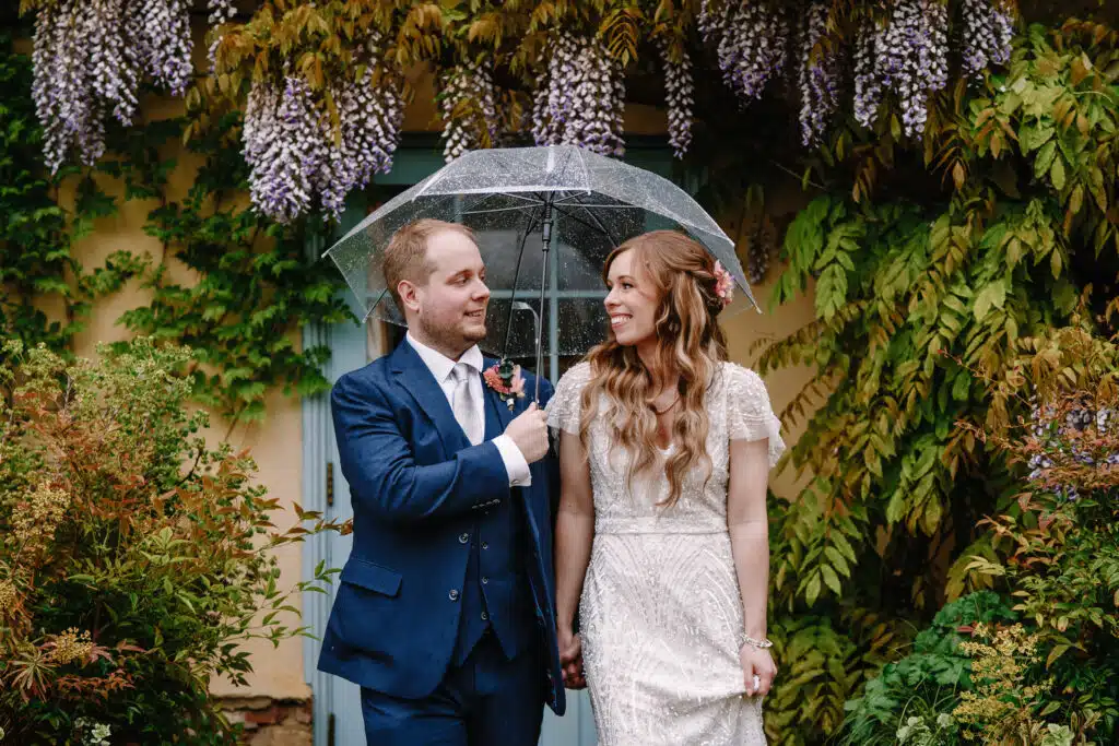 Bride and Groom standing under umbrella with gorgeous Wisteria above them at outdoor country wedding venue