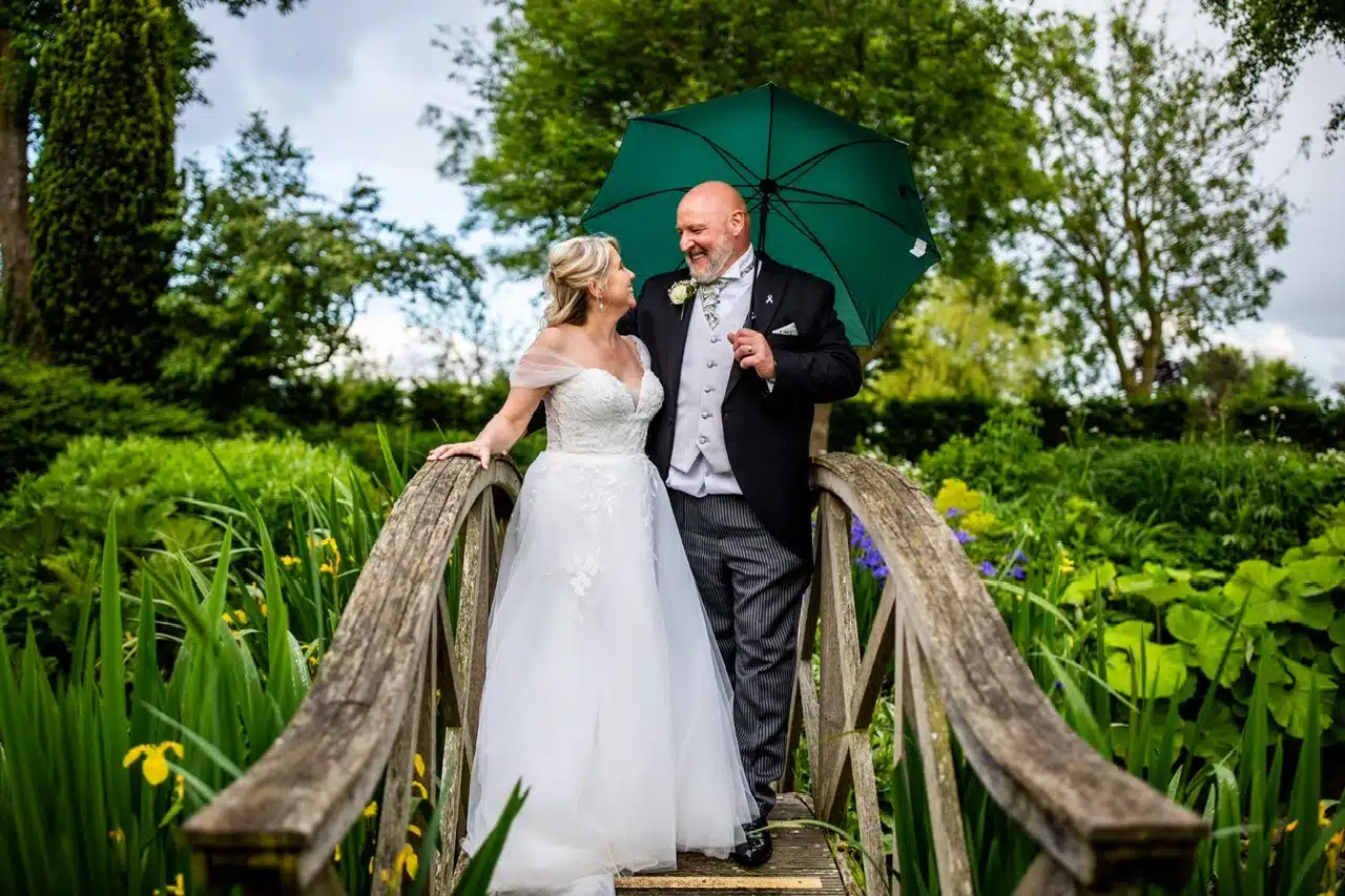 Bride and Groom under a green umbrella on the Monet Bridge surrounded by formal gardens at outdoor garden wedding venue