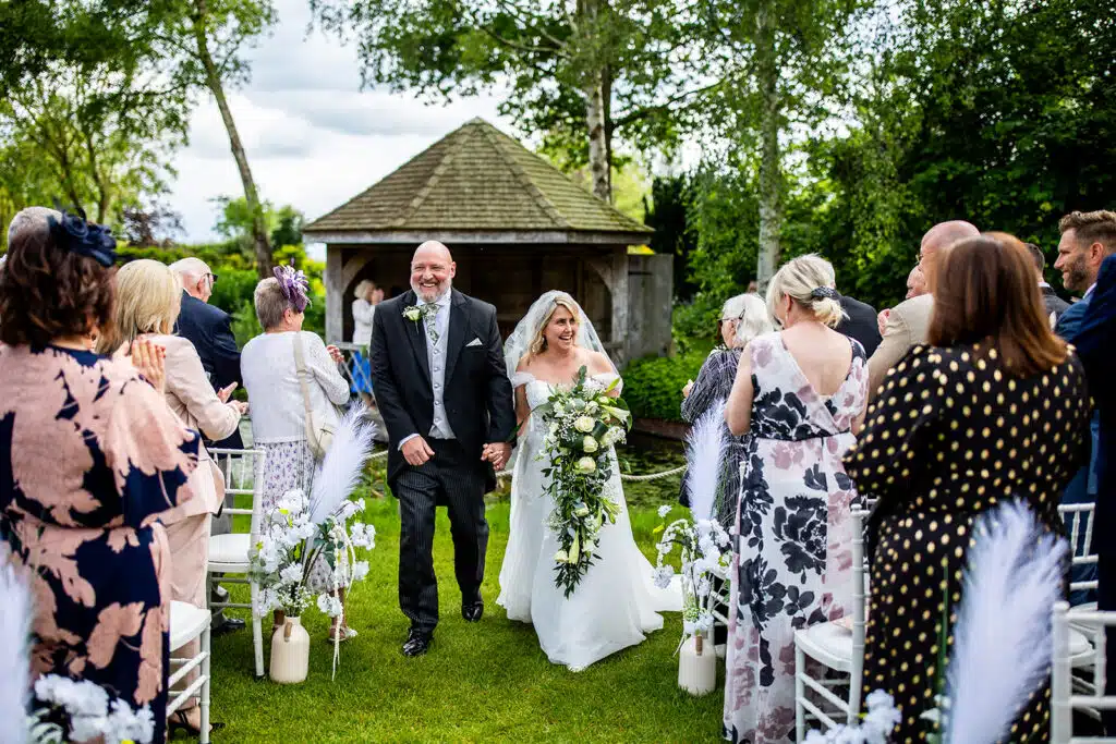 Bride and Groom walking up the aisle with the Summer House behind them at outdoor garden ceremony