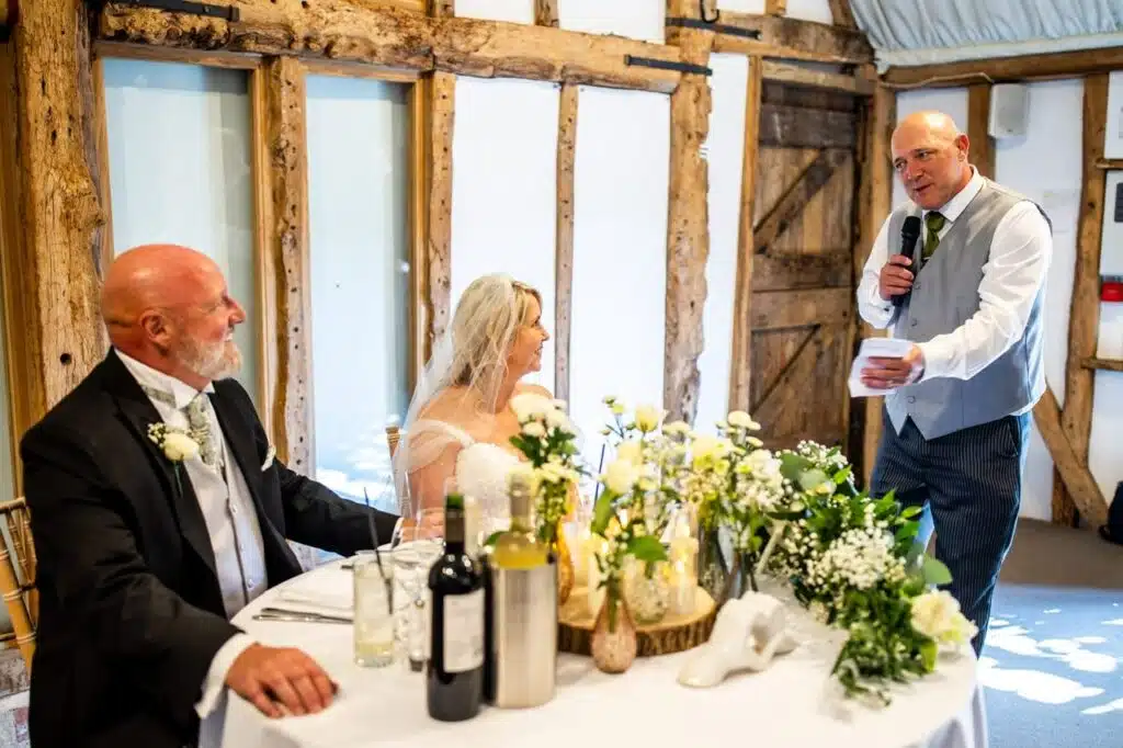 Speeches taking place in the rustic Tudor Barn at barn wedding venue. Bride and Groom sitting at sweetheart table