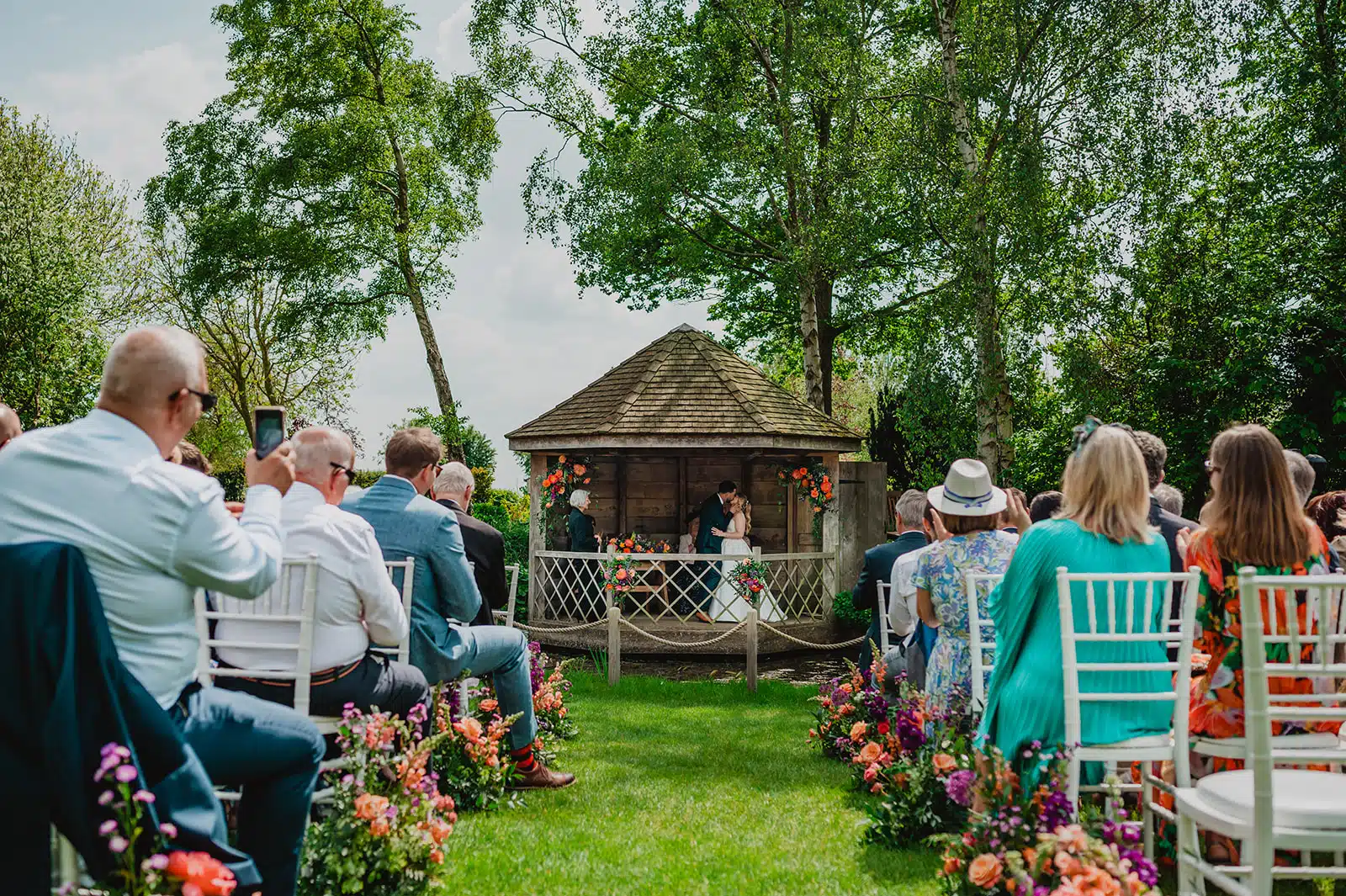 Bride and Groom in the Summer House saying their wedding vowels and having sharing a kiss amongst their friends and family with flowers down the aisle at barn wedding venue
