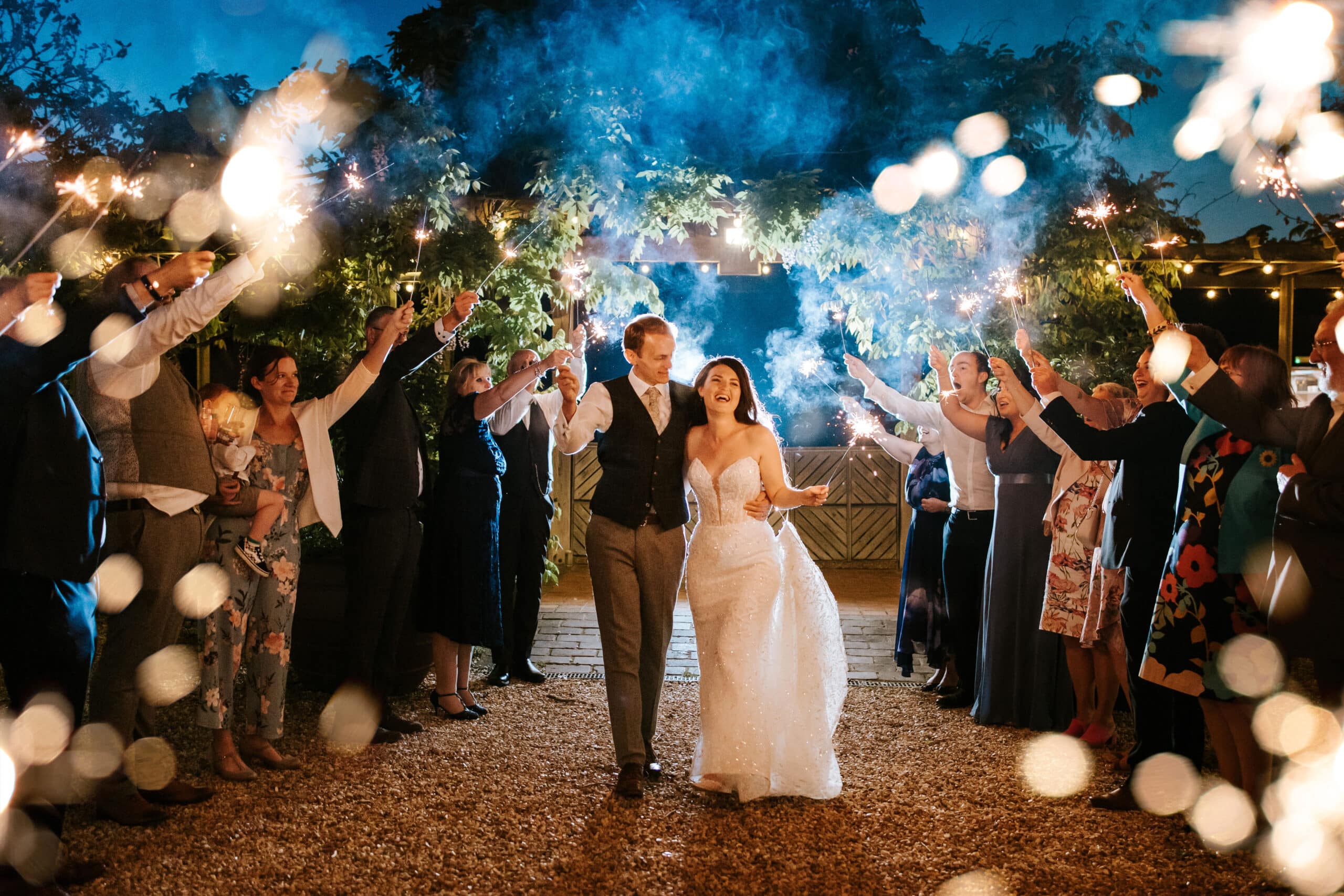 Sparklers at dusk at farm wedding venue in courtyard. Bride and groom share a moment with their guests holding sparklers high against a blue dusky sky