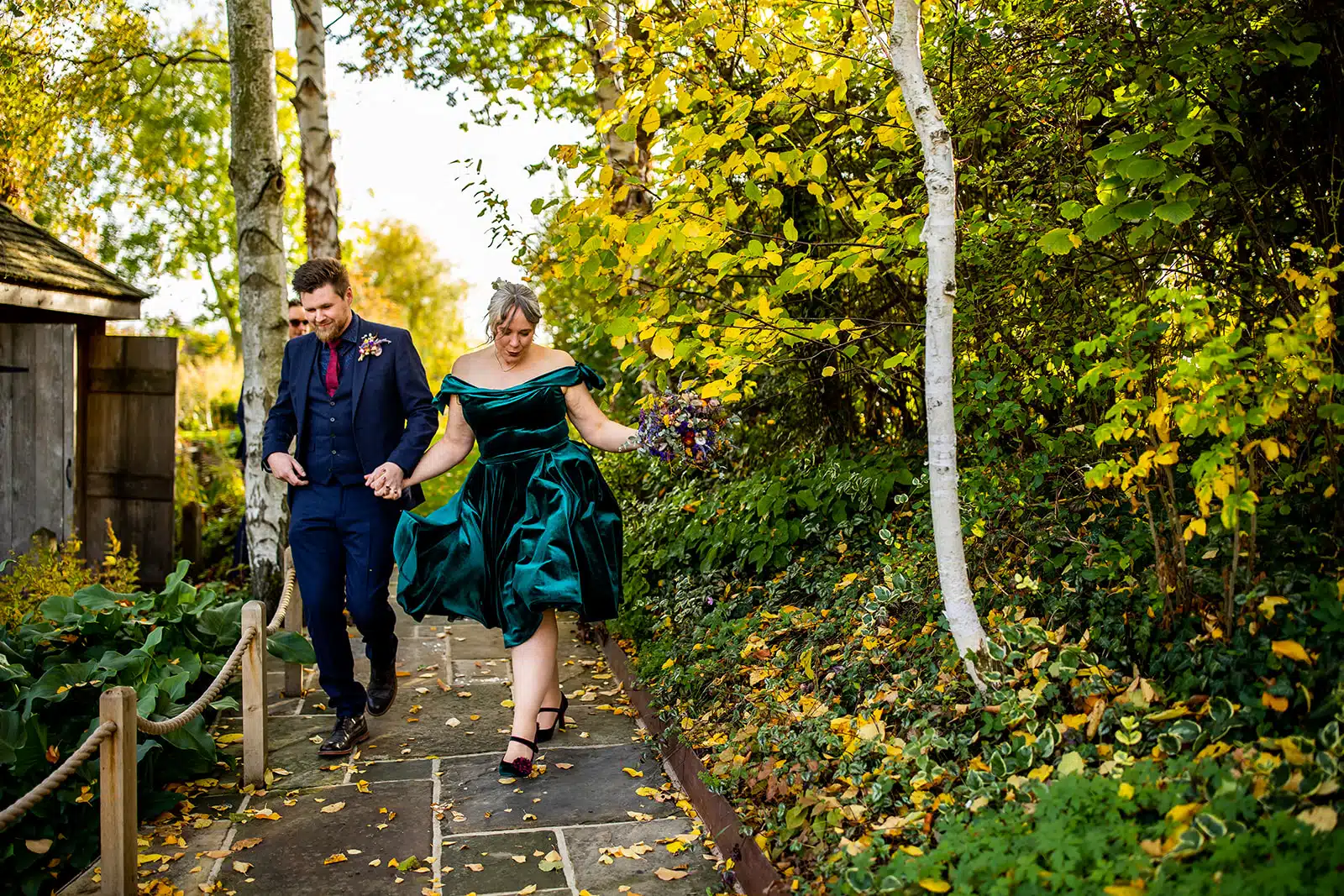 Bride wearing bold green dress walking out of the Summerhouse with stunning autumn colours in the formal gardens at barn wedding venue based in Hertfordshire