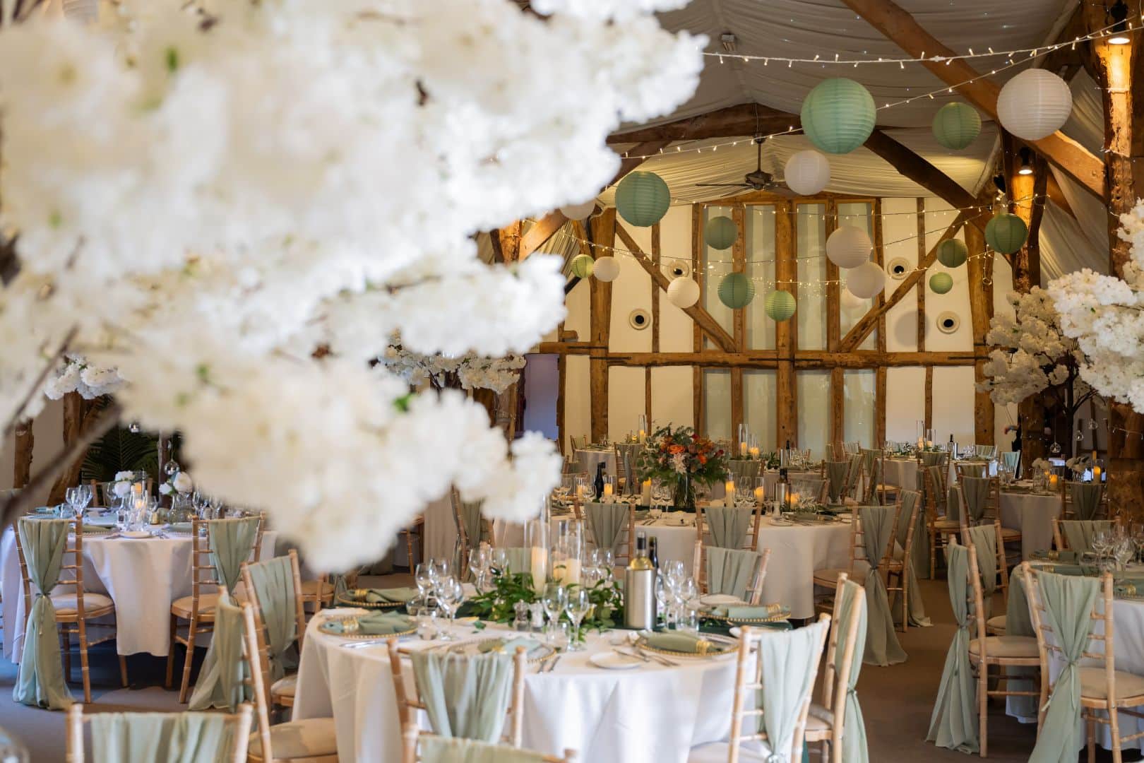 Beautiful sage green decor with white blossom trees on the tables in the rustic Tudor Barn at eco conscious wedding venue