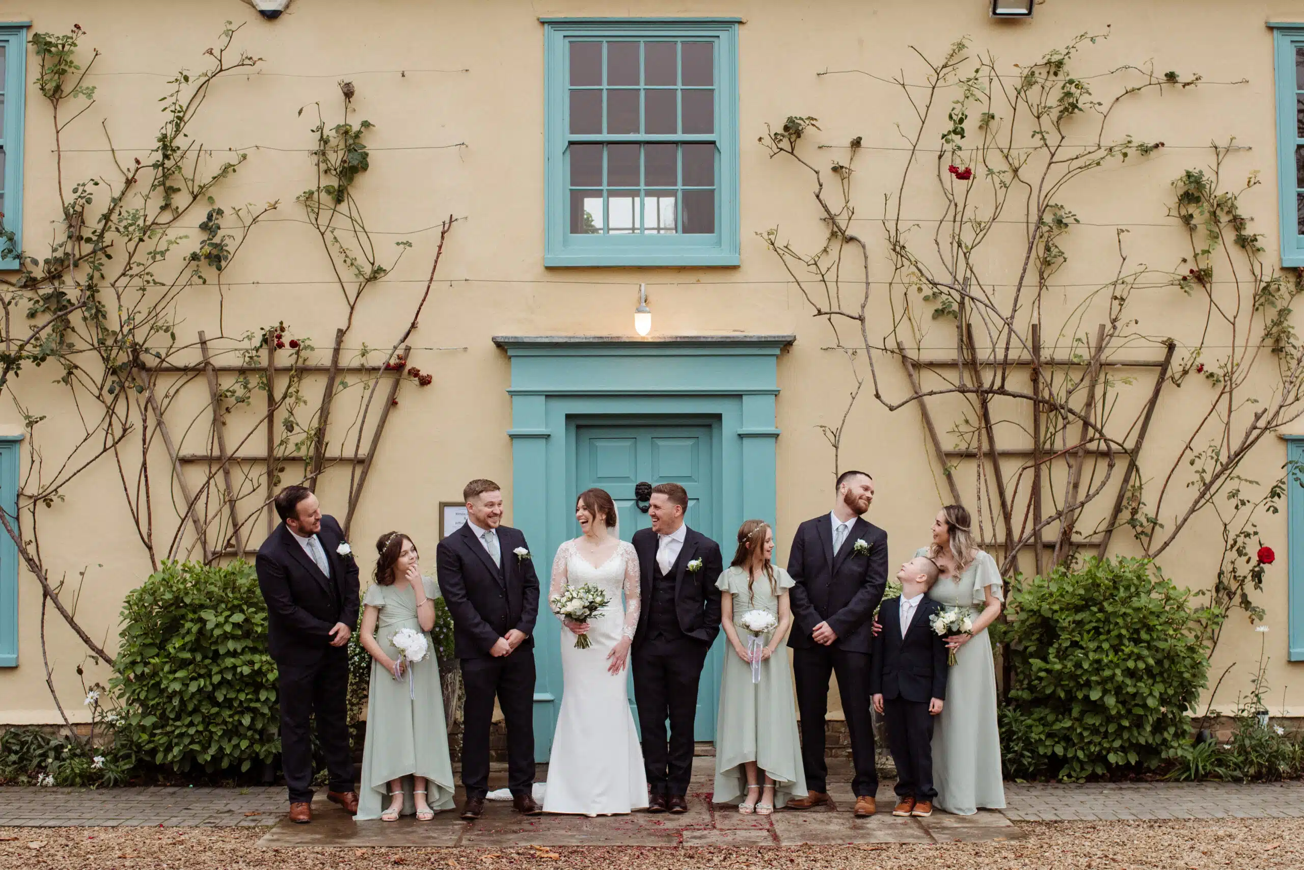 Bride and Groom standing with their bridal party at the front of the rose adorned Farmhouse at eco conscious wedding venue