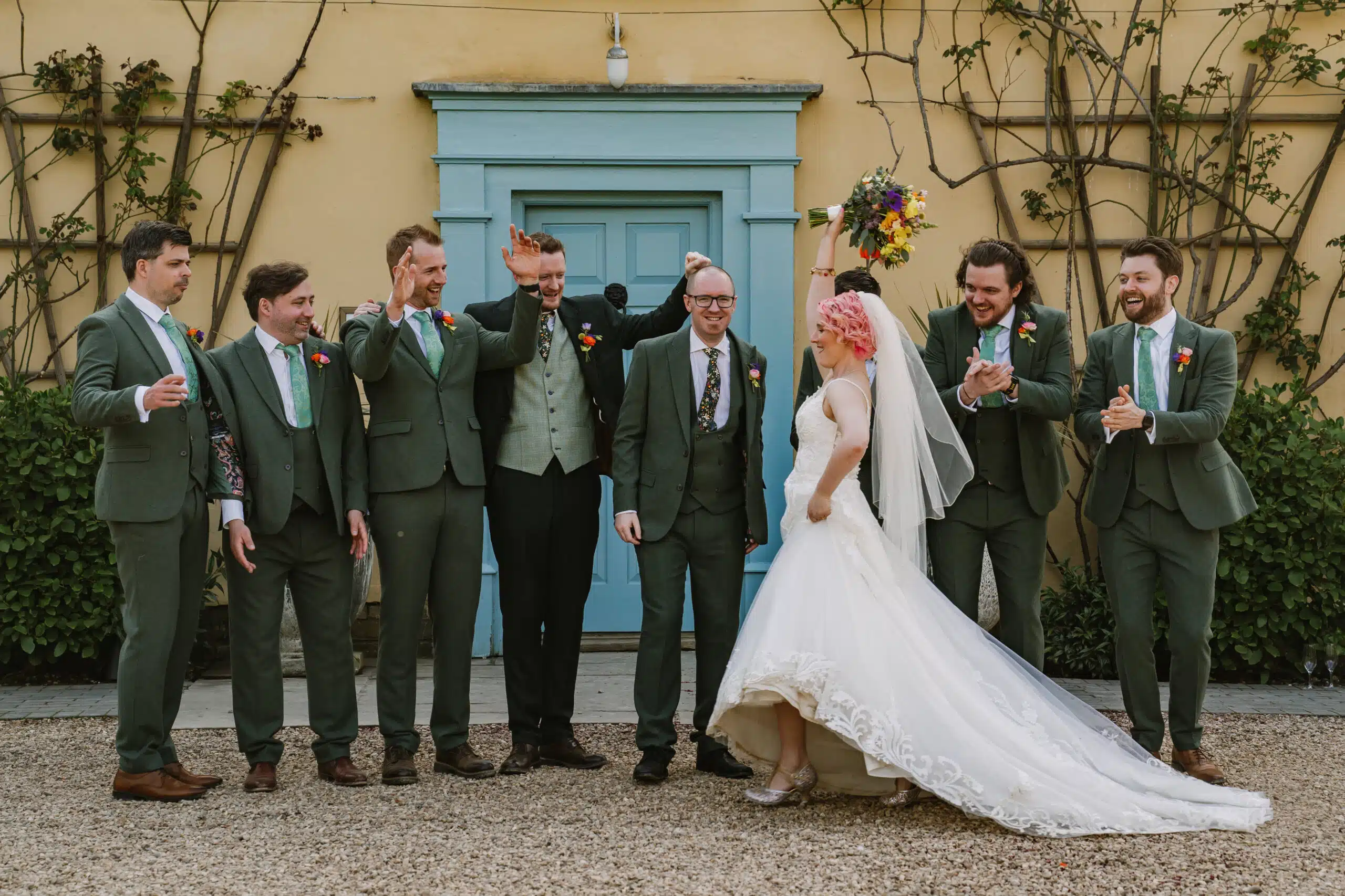 The Bride and the Groomsmen having a fun moment at the front of the charming farmhouse with blue door at eco conscious wedding venue near Cambridgeshire