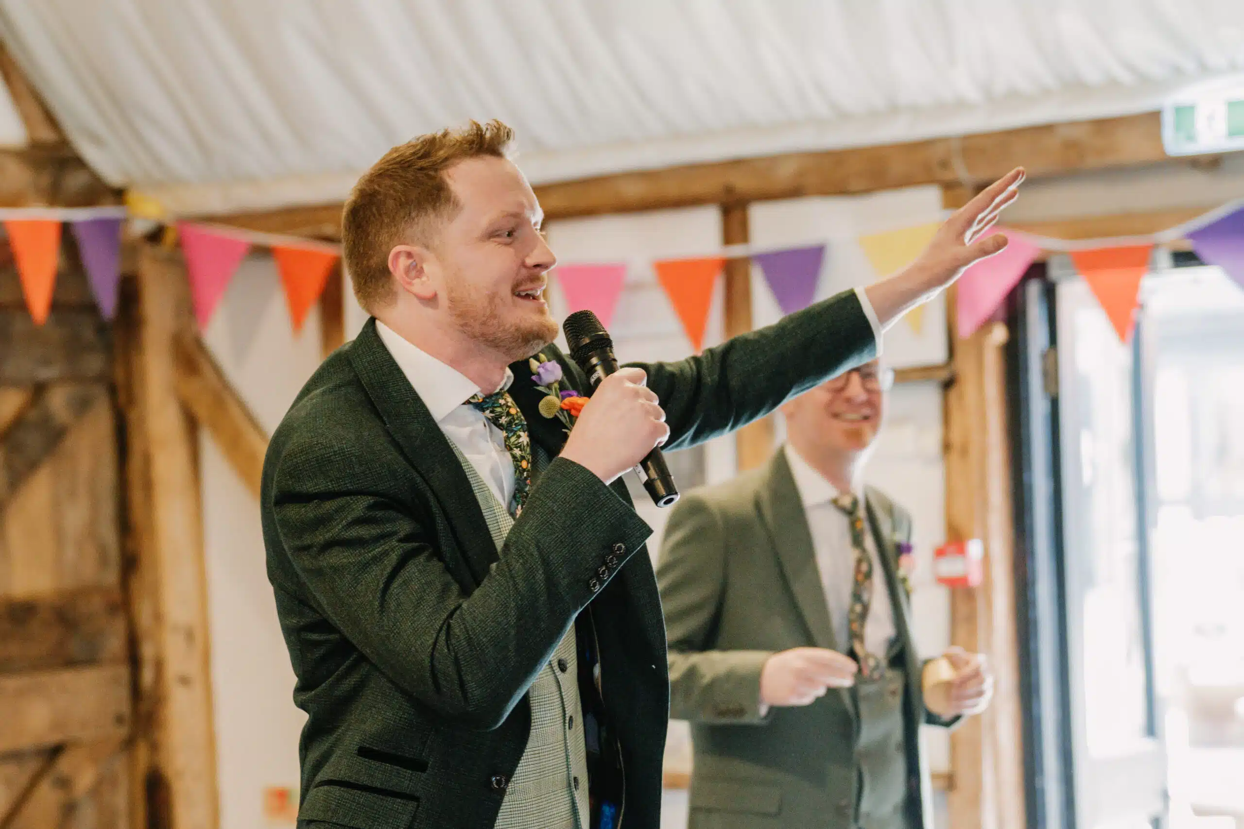 The Groomsmen in the Tudor Barn with mic in hand during the speeches with bright colourful bunting behind 