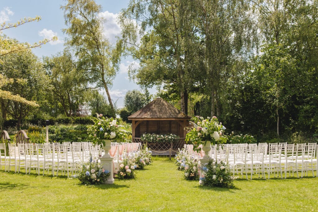Wedding ceremony set up in stunning wedding garden at countryside wedding venue with white ceremony chairs, a pretty garden summerhouse and beautiful flower arrangements