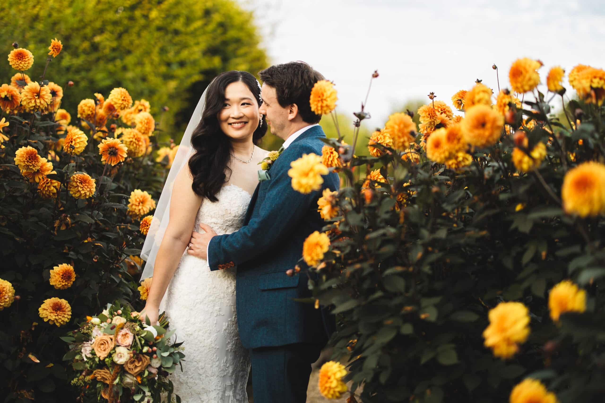 Bride and Groom standing in amongst the orange dahlia's at South Farm barn wedding venue with stunning seasonal gardens and grounds