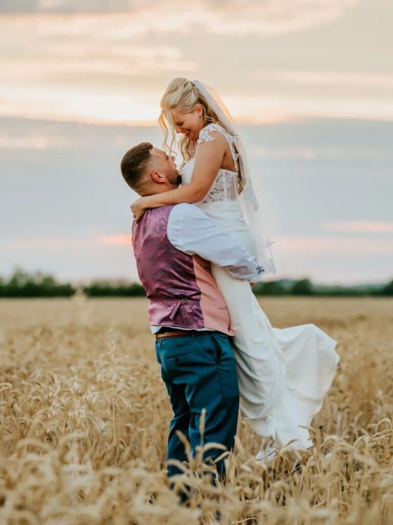 Groom lifts bride in a celebratory moment in a field of corn at their farm wedding with a stunning pink and blue sunset in background
