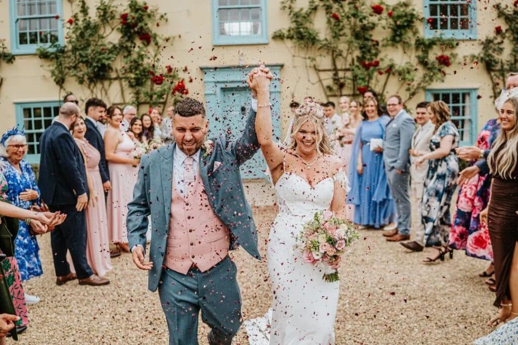 Bride and Groom celebrate their marriage under a rose petal confetti shower with their guests in front of a countryside farm house