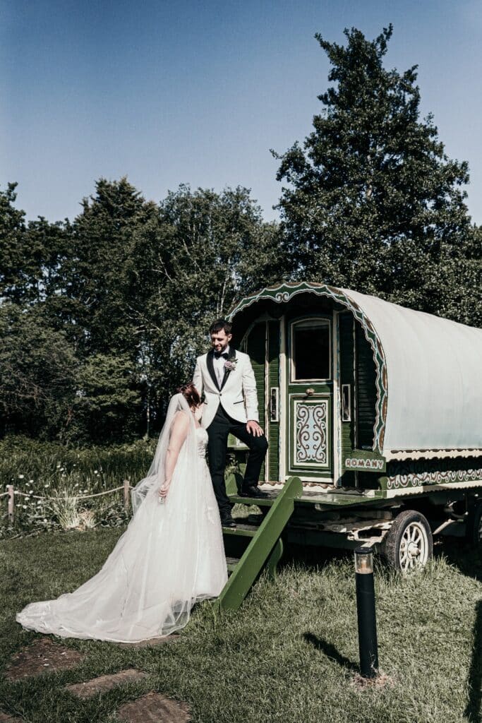 Bride and groom on wedding day in front of cream and green romany wagon at boho wedding venue