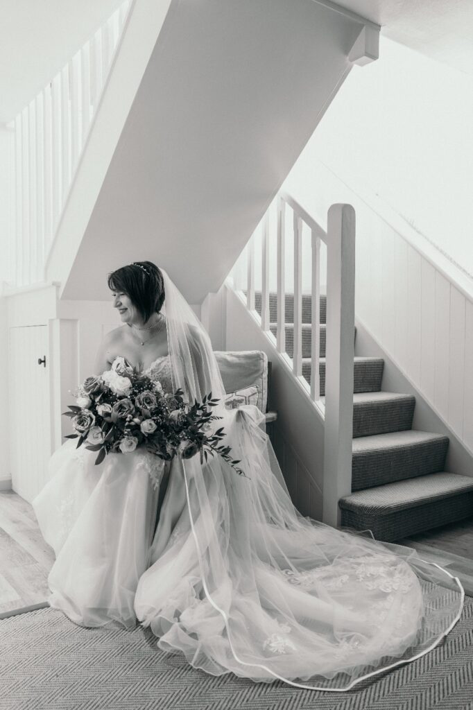 Black and white photo of Bride in stunning wedding dress with large bouquet seated beneath staircase at barn wedding venue