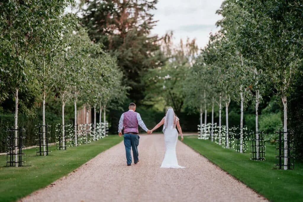 Bride and groom walking away from the camera down tree lined driveway with twinkly fairy lights on their summer wedding day